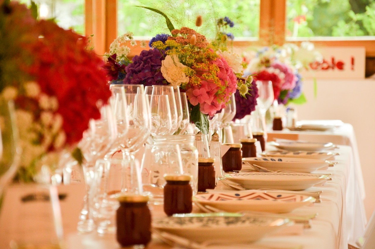Table dressée avec vaisselle élégante, verres à pied, pots de confiture et bouquets colorés dans un cadre lumineux.