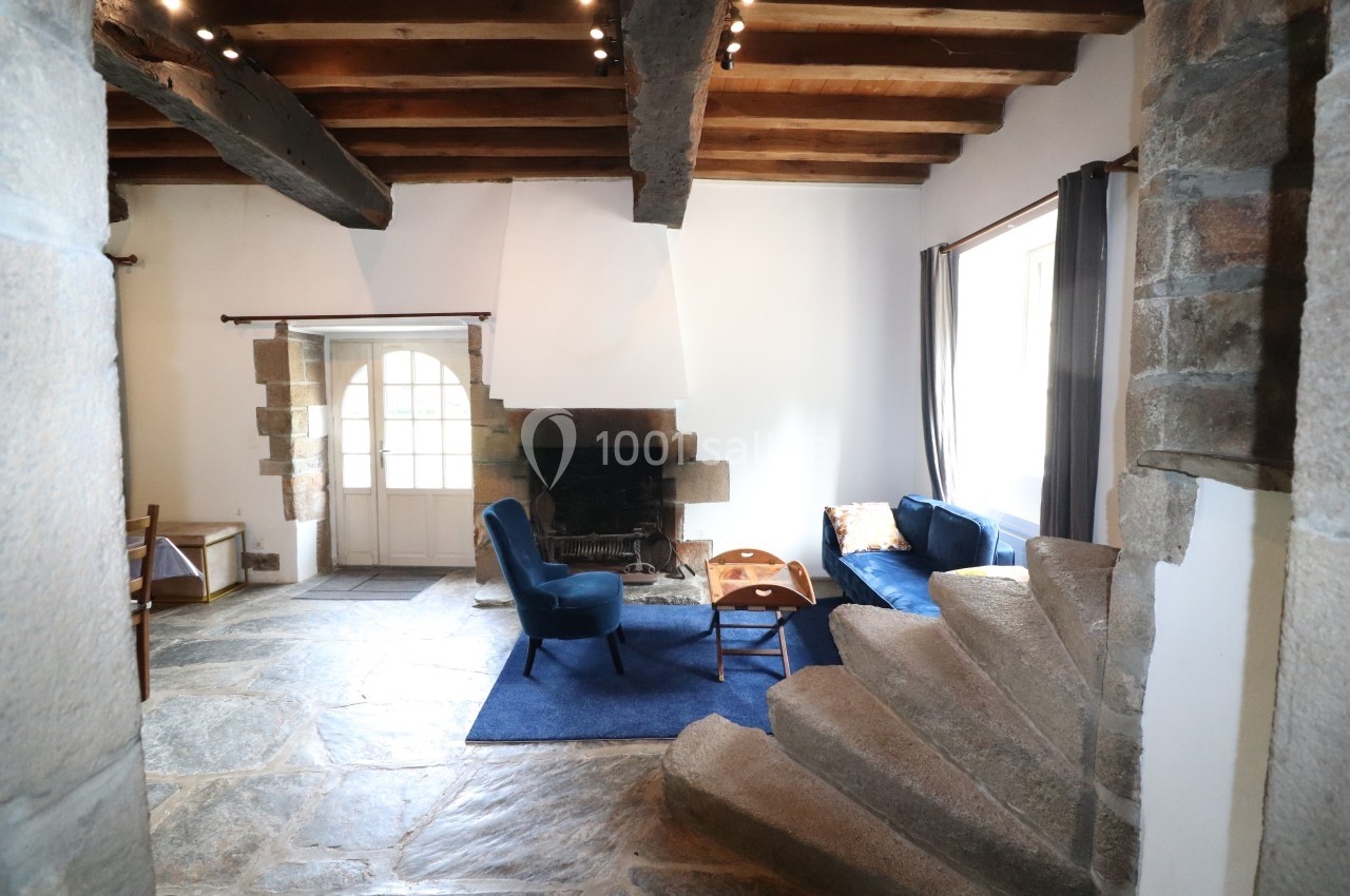 Salon avec cheminée, poutres apparentes, escalier en pierre, fauteuils bleus et lumière naturelle entrant par les fenêtres.
