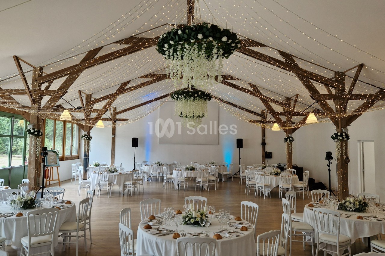 Salle de réception décorée avec des guirlandes lumineuses, tables rondes dressées et poutres apparentes en bois.