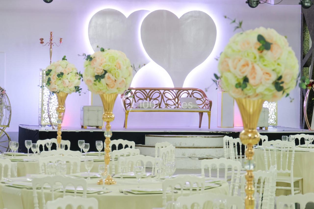 Salle de réception décorée avec des tables dressées, des bouquets de fleurs et une scène ornée de deux cœurs lumineux.