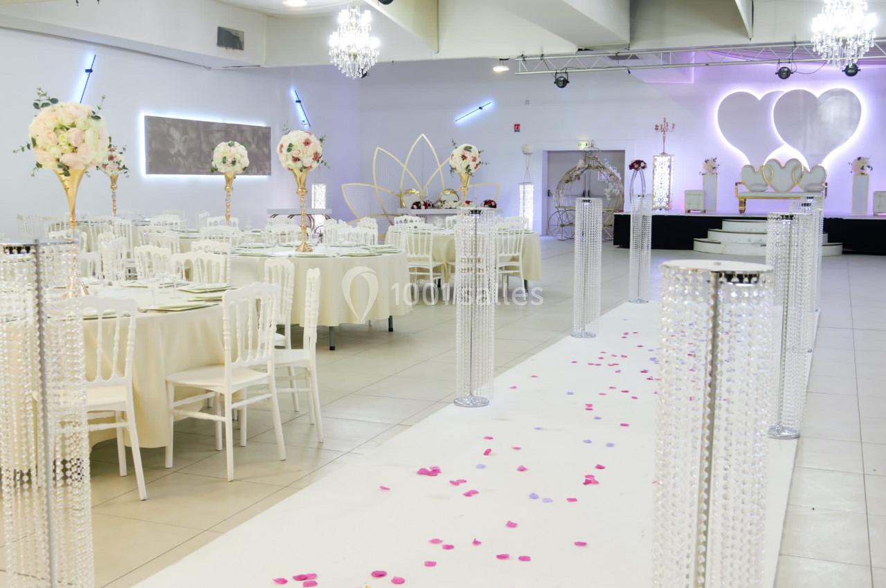 Salle de réception décorée pour un mariage avec tables dressées, chandeliers, pétales au sol et estrade en arrière-plan.