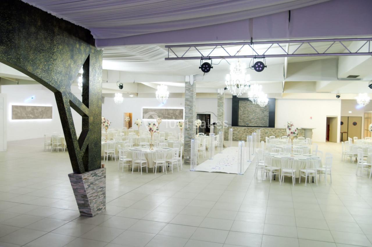 Salle de réception spacieuse avec tables rondes dressées, décorations florales et éclairage élégant.
