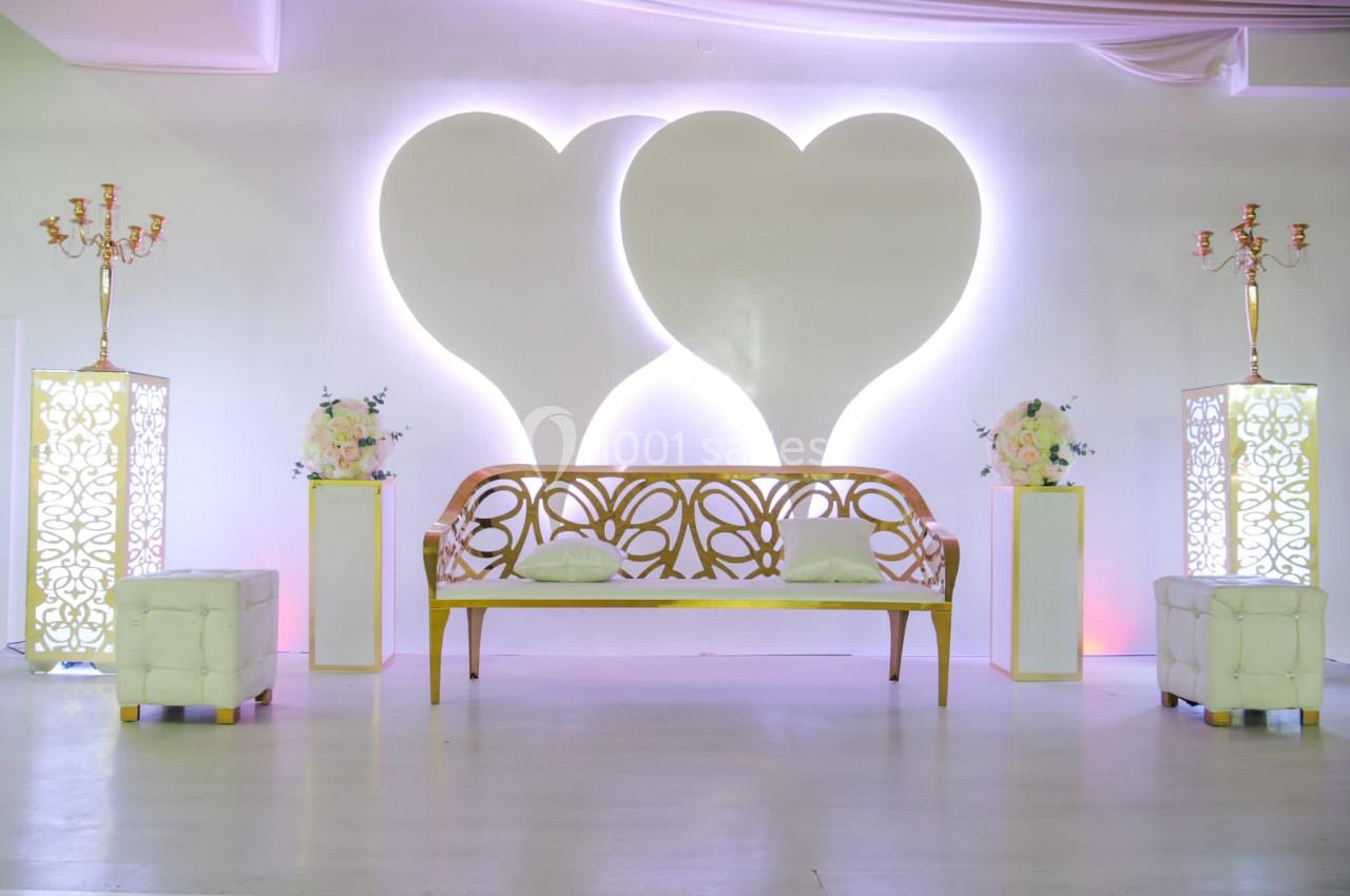 Banc décoratif doré devant un mur blanc orné de deux cœurs lumineux, entouré de bougeoirs et de fleurs.