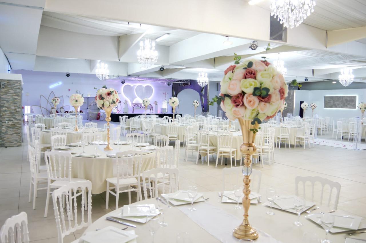 Salle de réception décorée avec des tables rondes, nappes blanches, chaises blanches et centres de table fleuris.