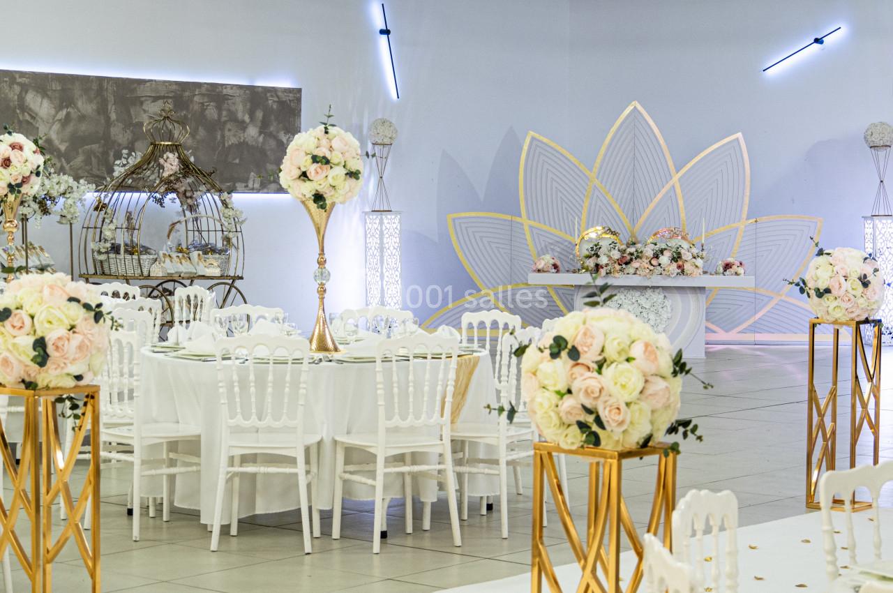 Salle de réception décorée avec des tables blanches, des compositions florales et un arrière-plan orné d'un motif floral…