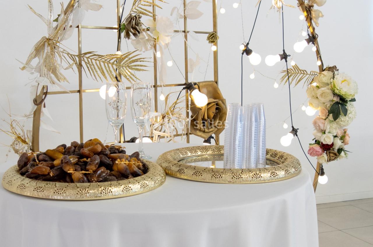 Décoration de table avec structure dorée ornée de fleurs, guirlandes lumineuses et plateaux contenant des dattes.
