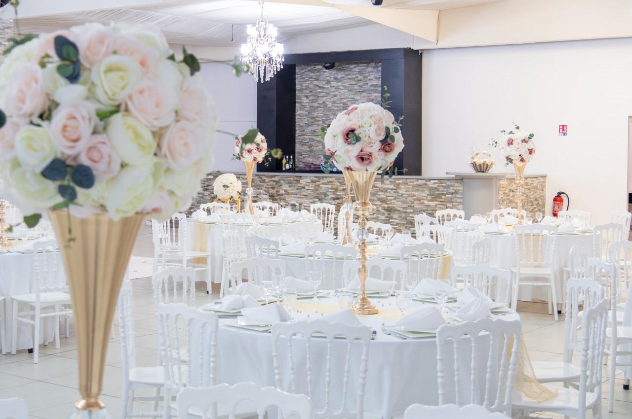 Salle de réception décorée avec des tables rondes, nappes blanches, chaises assorties et centres de table fleuris.