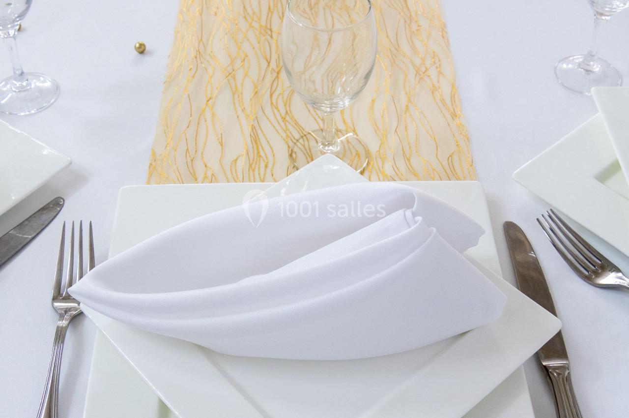 Table dressée avec une nappe blanche, un chemin de table doré, des assiettes carrées et des serviettes pliées.