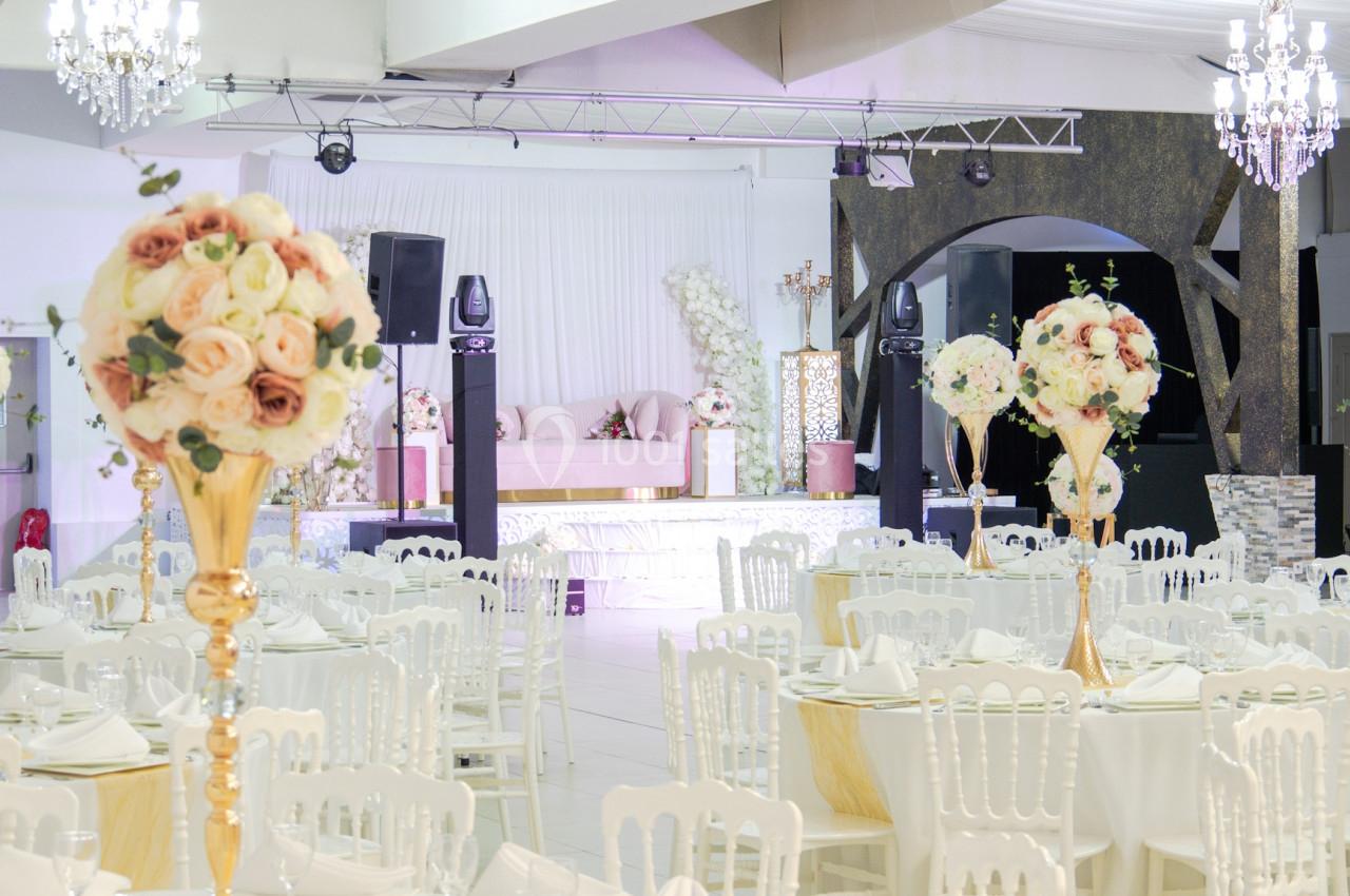Salle de réception élégante décorée de tables rondes, chaises blanches et centres de table fleuris dans des tons pastel.