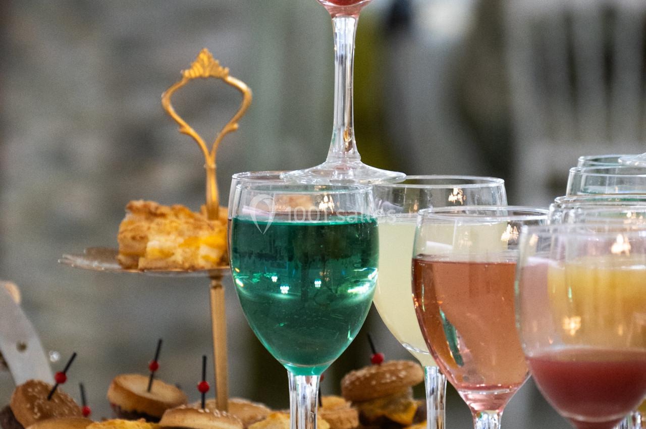 Verres de cocktails colorés sur une table, accompagnés d'un plateau de petits fours en arrière-plan flou.