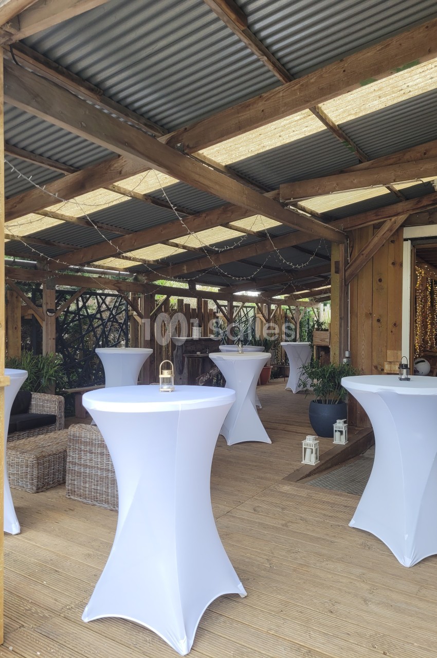 Salle en bois avec tables hautes recouvertes de nappes blanches, décorée de plantes et guirlandes lumineuses.