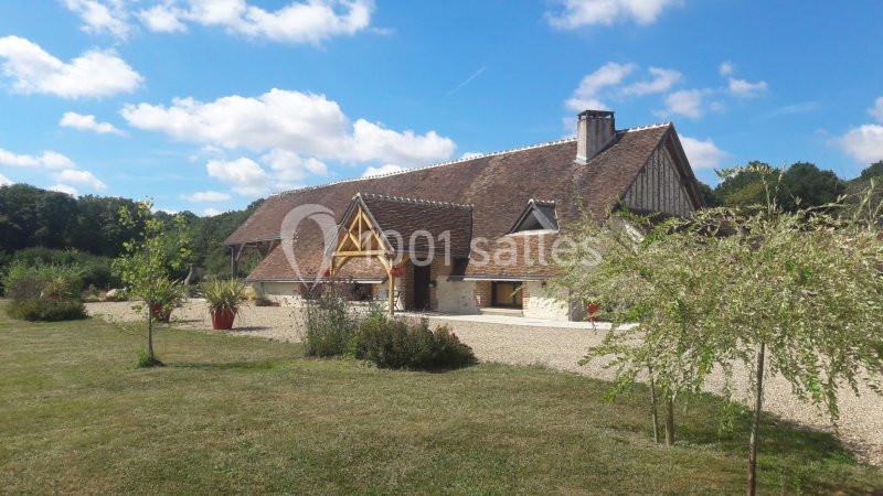 Bâtiment rural en pierre et bois avec toit en tuiles, entouré de pelouse, arbres et ciel dégagé.