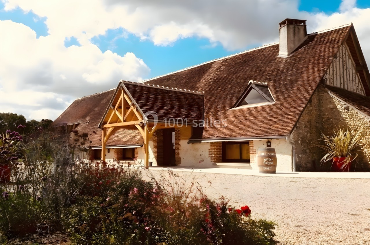 Façade d'une maison traditionnelle en pierre avec toit en tuiles, entourée de fleurs et d'un ciel partiellement nuageux.