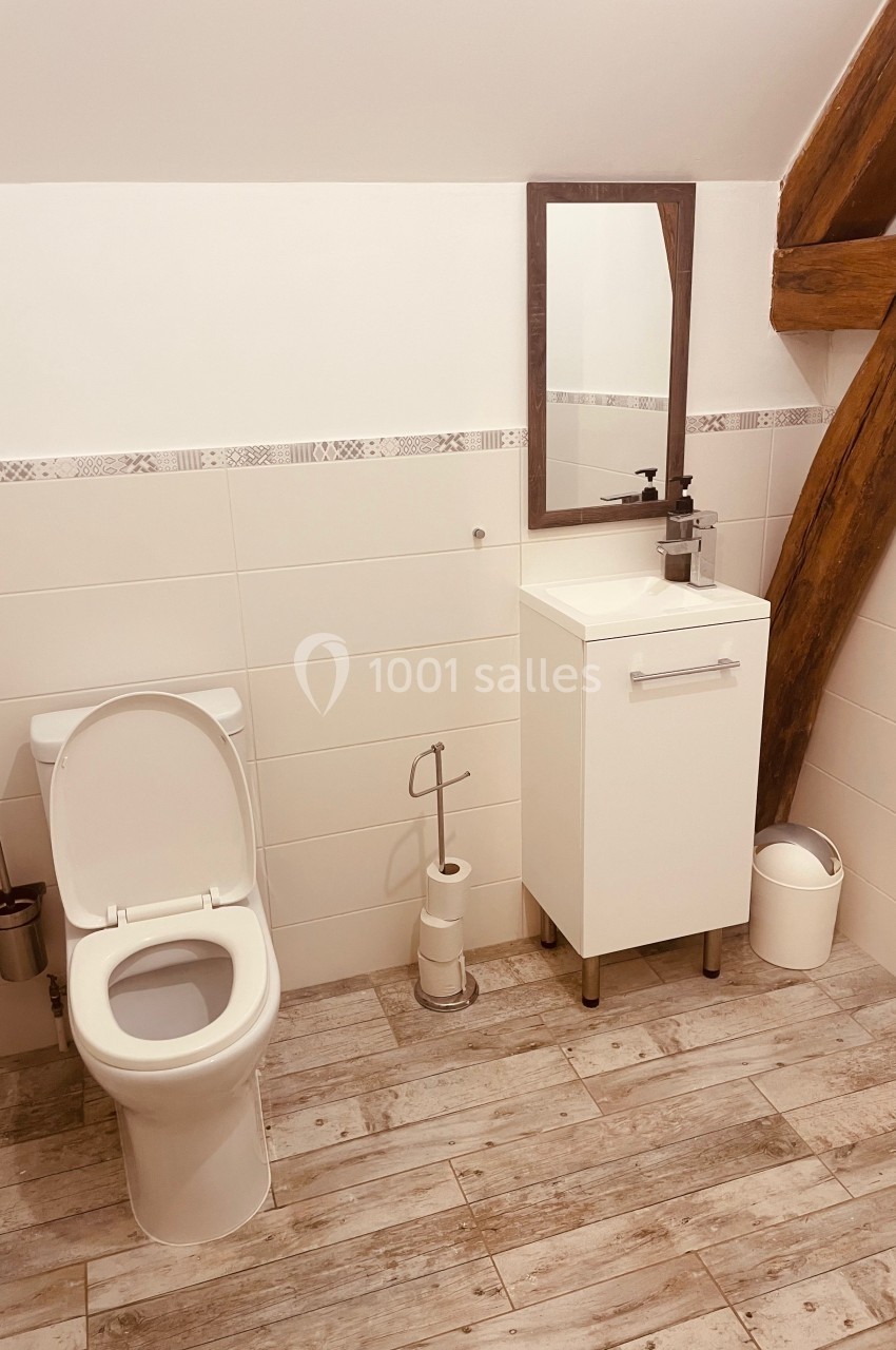 Toilettes dans une salle de bain lumineuse avec sol en bois, meuble blanc, miroir et poutre apparente.