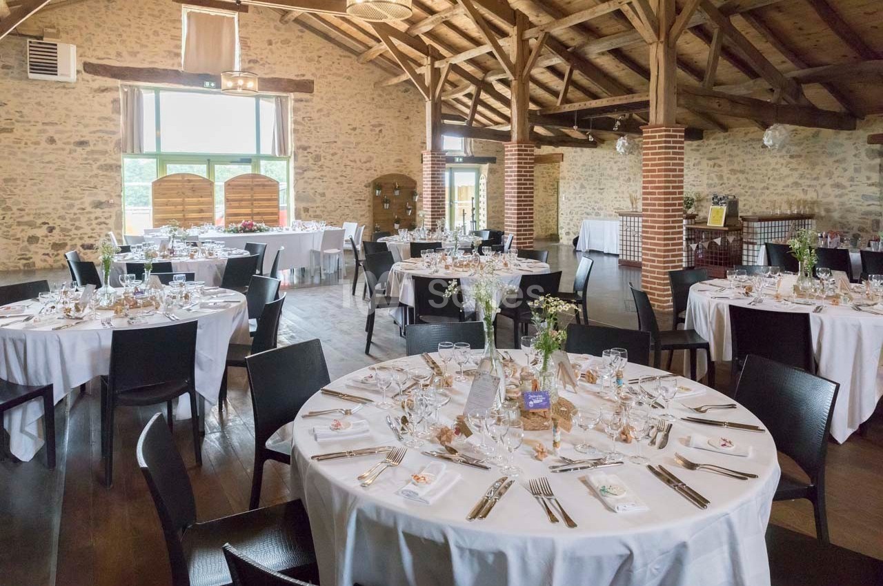 Salle de réception avec tables rondes dressées pour un événement, décor rustique avec murs en pierre et poutres apparentes.