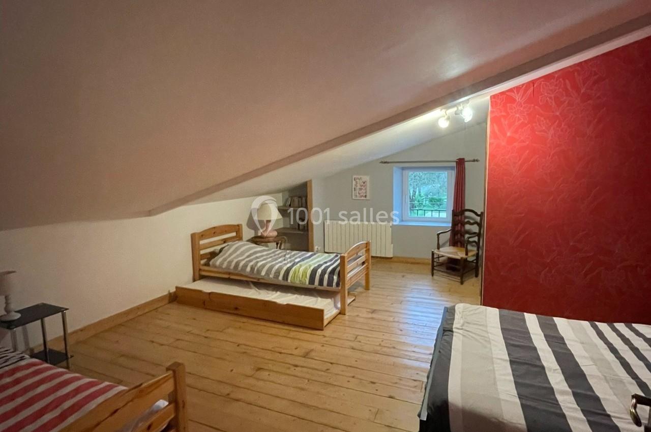 Chambre mansardée avec trois lits simples, parquet en bois, mur rouge décoratif et fenêtre laissant entrer la lumière…