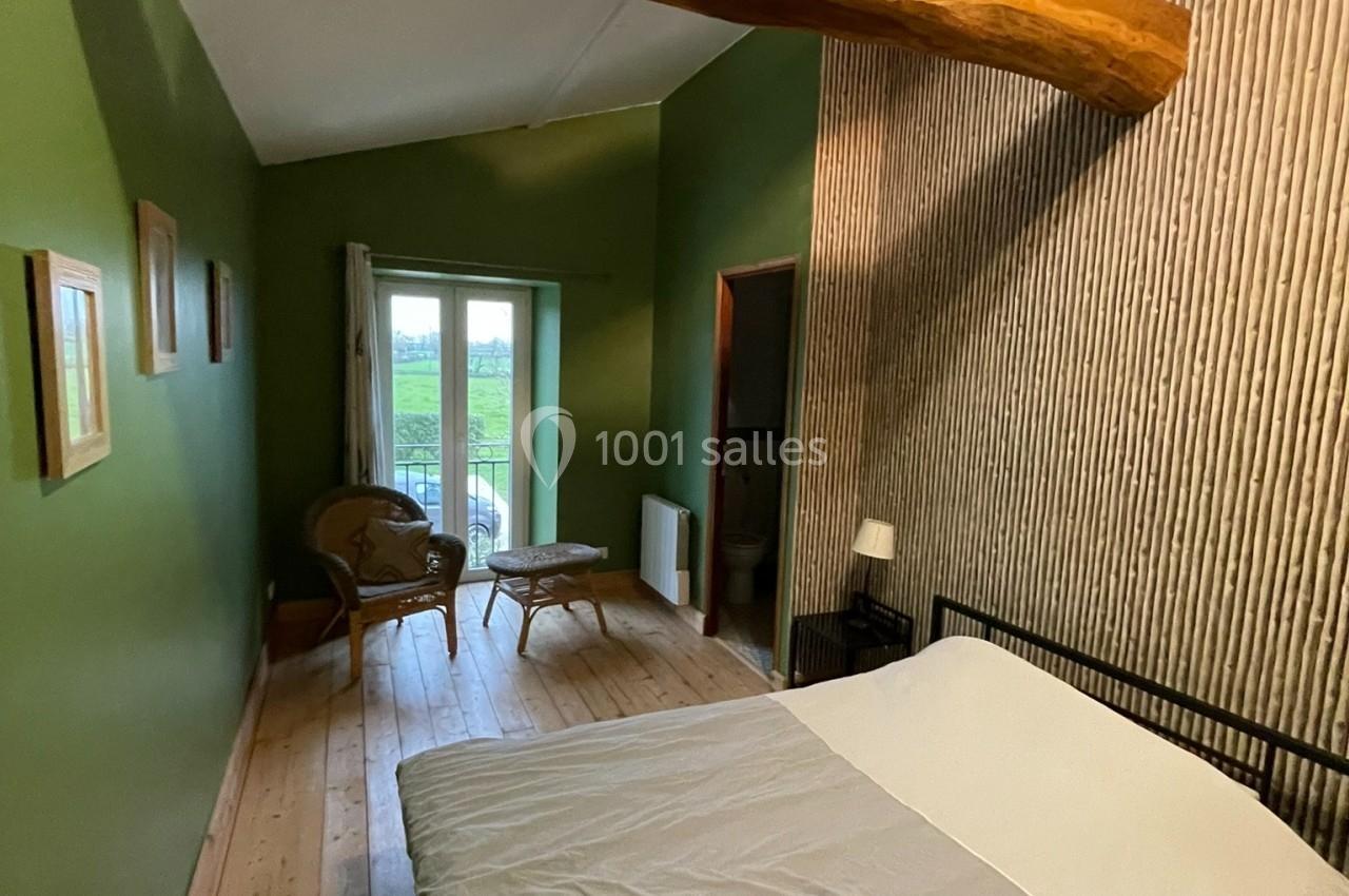 Chambre avec murs verts, poutre en bois apparente, lit double, parquet et vue sur l'extérieur par une porte-fenêtre.