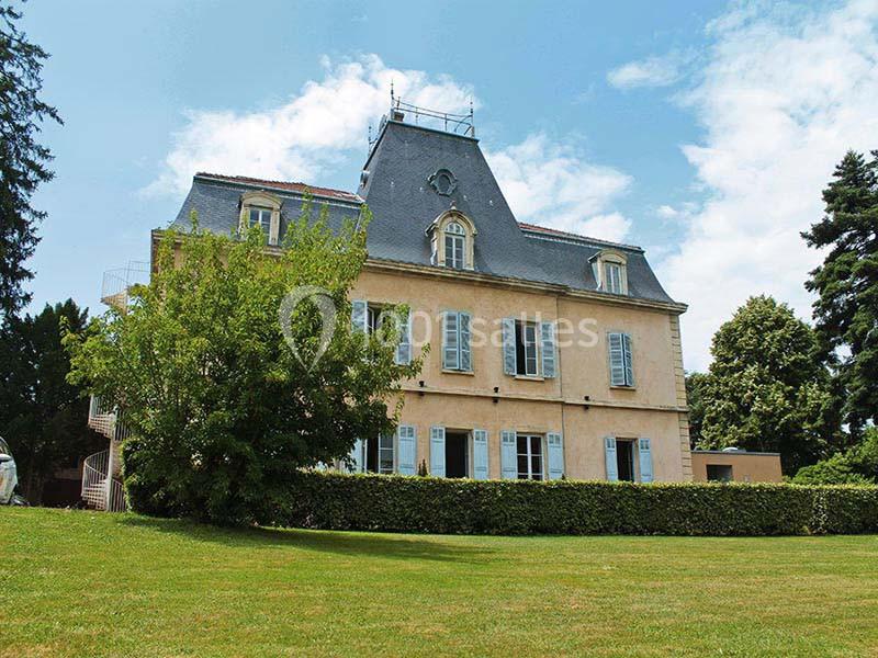 Grande maison ancienne avec toit en ardoise, entourée de pelouse, d'arbres et d'un ciel partiellement nuageux.