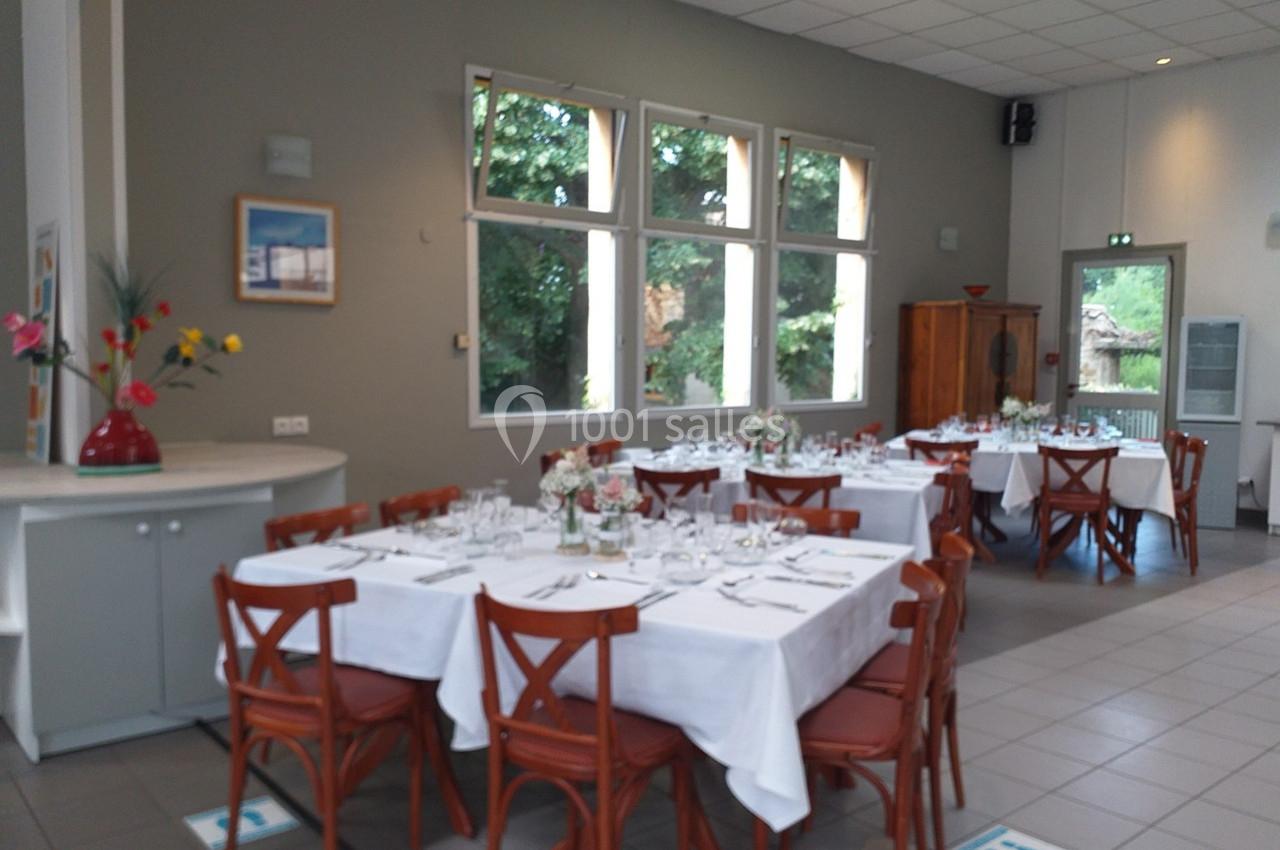 Salle lumineuse avec tables dressées, nappes blanches, chaises en bois et grandes fenêtres donnant sur un jardin.