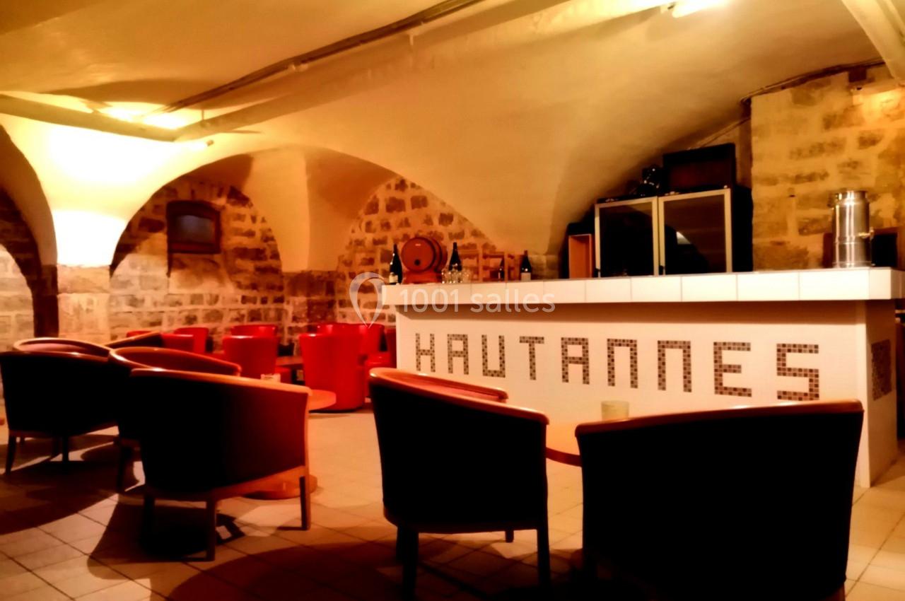 Salle voûtée avec des murs en pierre, un bar blanc avec l'inscription ’Hautannes’ et des fauteuils rouges.