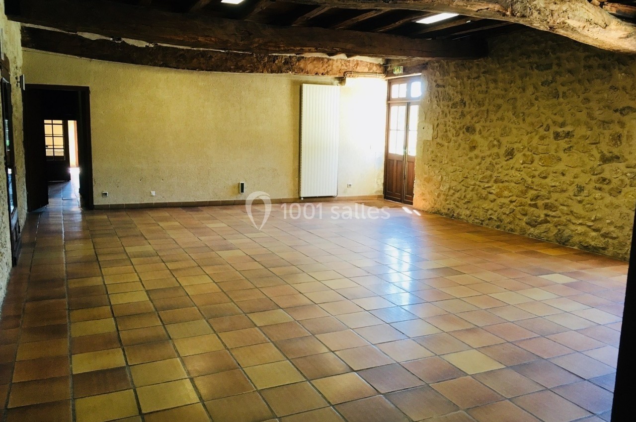 Salle vide avec sol en carrelage, murs en pierre et poutres apparentes, éclairée par une porte vitrée.