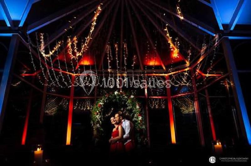 Un couple pose sous une arche décorée de feuillage et de lumières, dans un espace illuminé aux tons chauds et colorés.