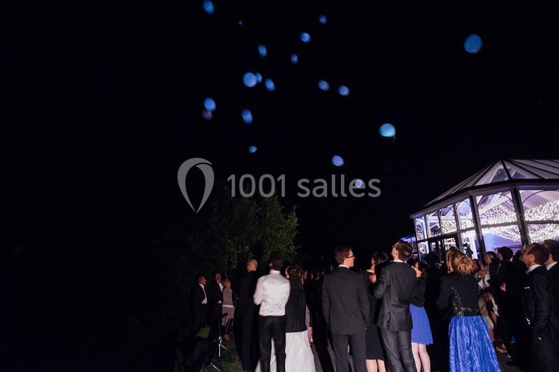 Un groupe de personnes en tenue de soirée observe des ballons lumineux s'élevant dans le ciel nocturne près d'une verrière…