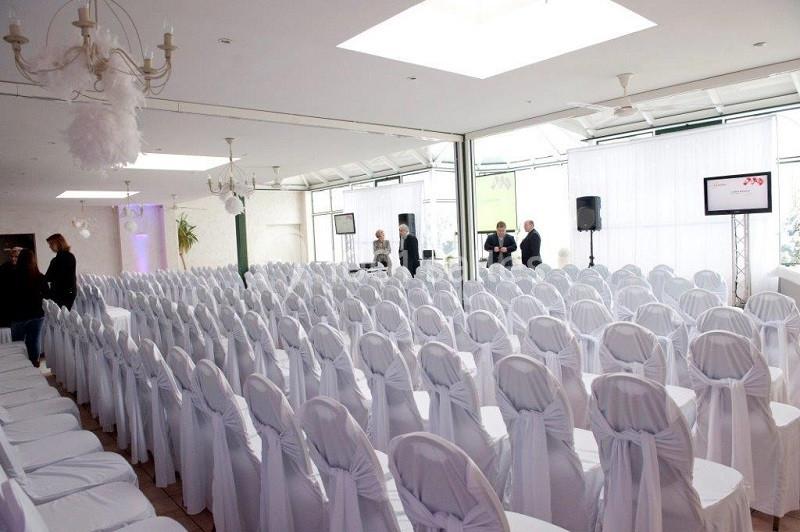 Salle de réception lumineuse avec des rangées de chaises blanches décorées et quelques personnes en arrière-plan.