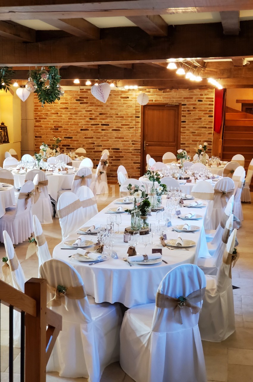 Salle de réception décorée pour un mariage, avec tables rondes dressées, chaises blanches et éclairage chaleureux.