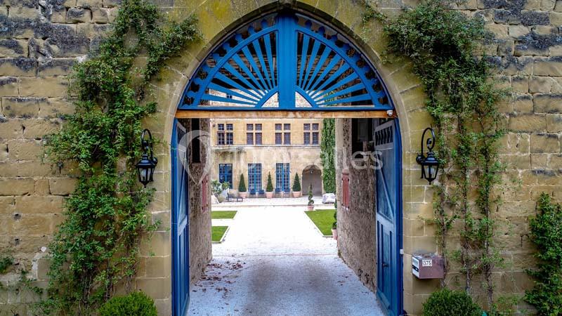 Porte en arc bleu encadrée de lierre, donnant sur une cour pavée avec un bâtiment en pierre en arrière-plan.
