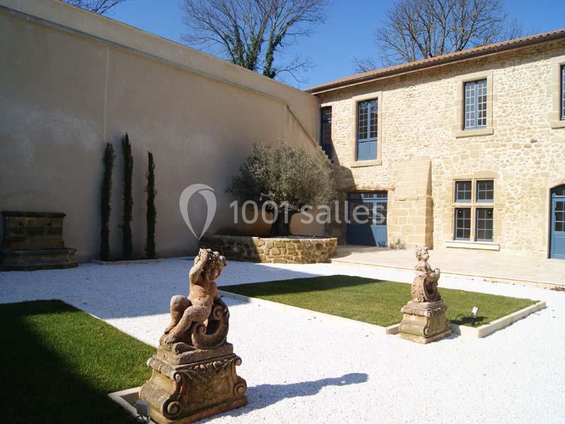 Cour intérieure avec pelouses, statues décoratives, arbre central et bâtiment en pierre aux volets bleus.