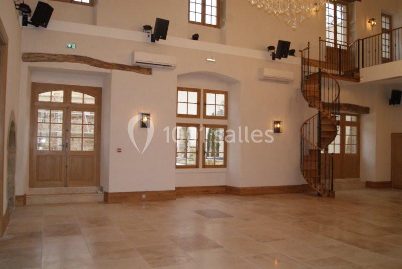 Salle lumineuse avec sol en pierre, escalier en colimaçon en bois, grandes fenêtres et portes vitrées.