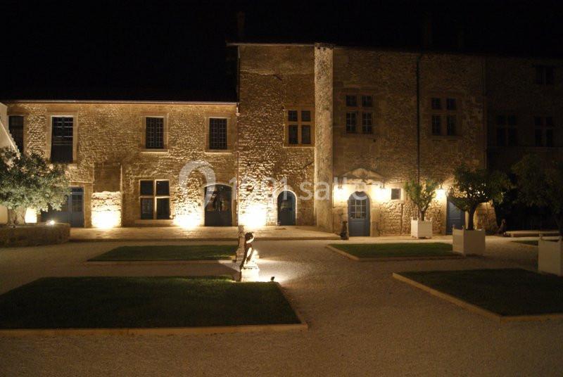 Façade en pierre d'un bâtiment historique éclairé la nuit, entourée de pelouses et d'allées gravillonnées.