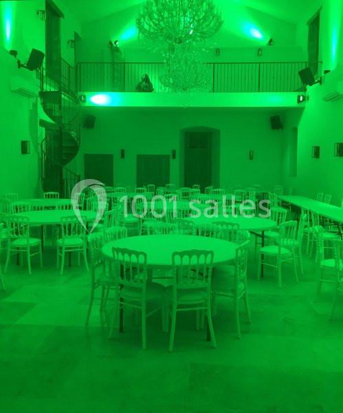 Salle de réception éclairée en vert, avec des tables rondes et des chaises blanches disposées sur un sol en pierre.