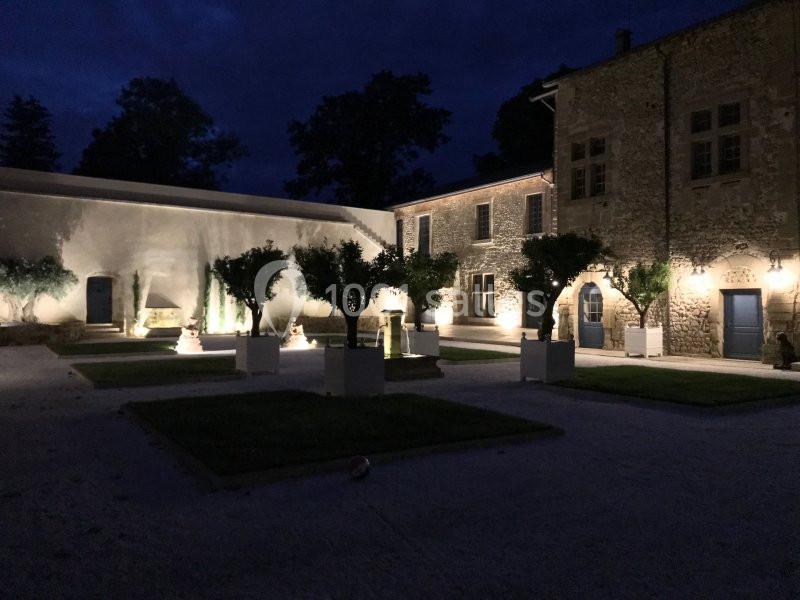 Cour intérieure d'un bâtiment en pierre éclairée par des lumières nocturnes, avec des arbres en pots et des pelouses.