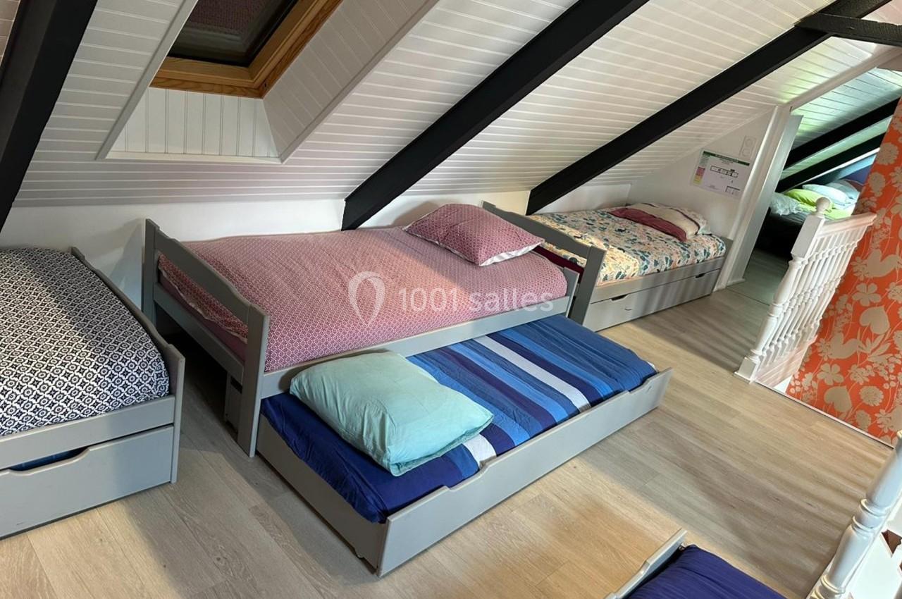 Chambre mansardée avec plusieurs lits simples, dont un avec tiroir-lit, parquet clair et mur partiellement tapissé.