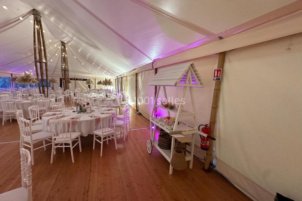 Salle de réception sous chapiteau avec tables rondes dressées, éclairage chaleureux et charrette décorative.