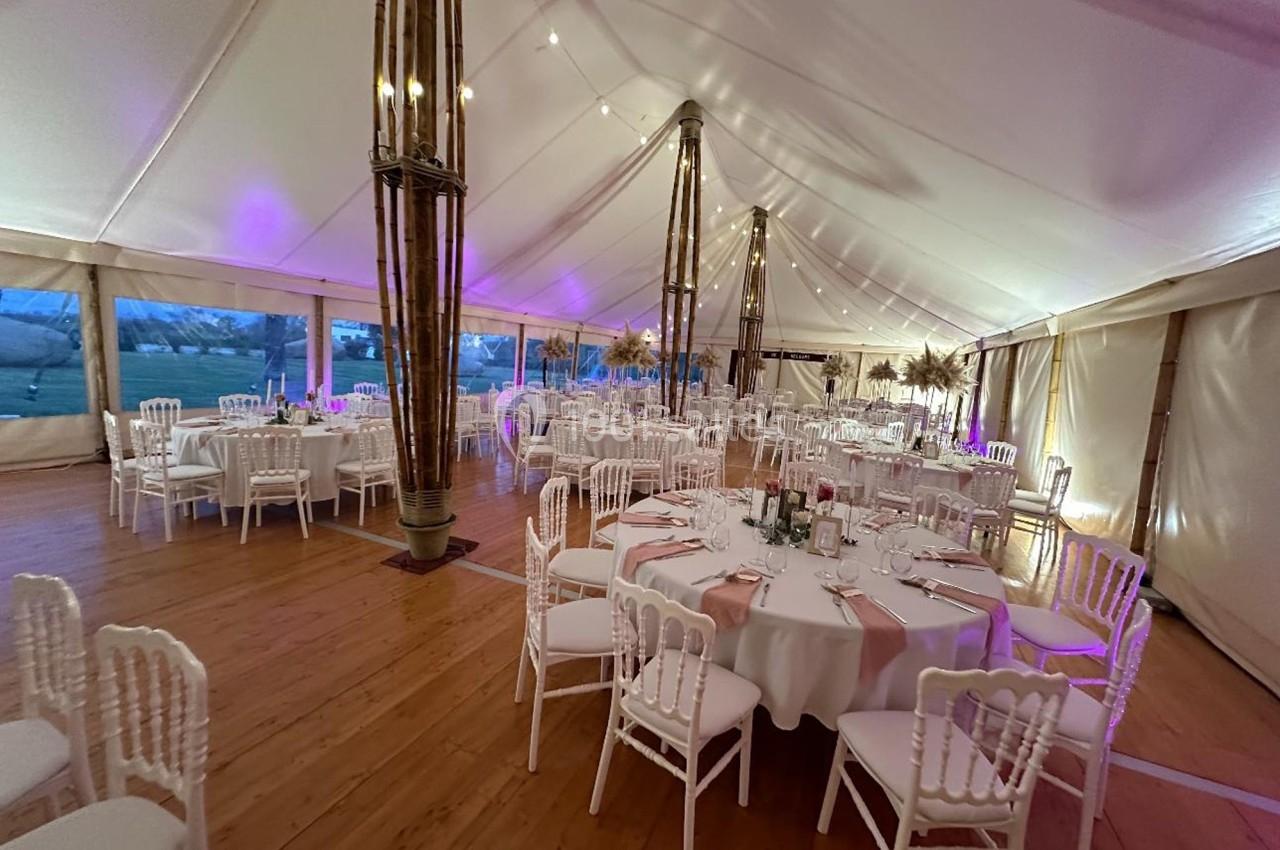 Salle de réception sous une tente avec tables rondes dressées, chaises blanches et éclairage chaleureux.