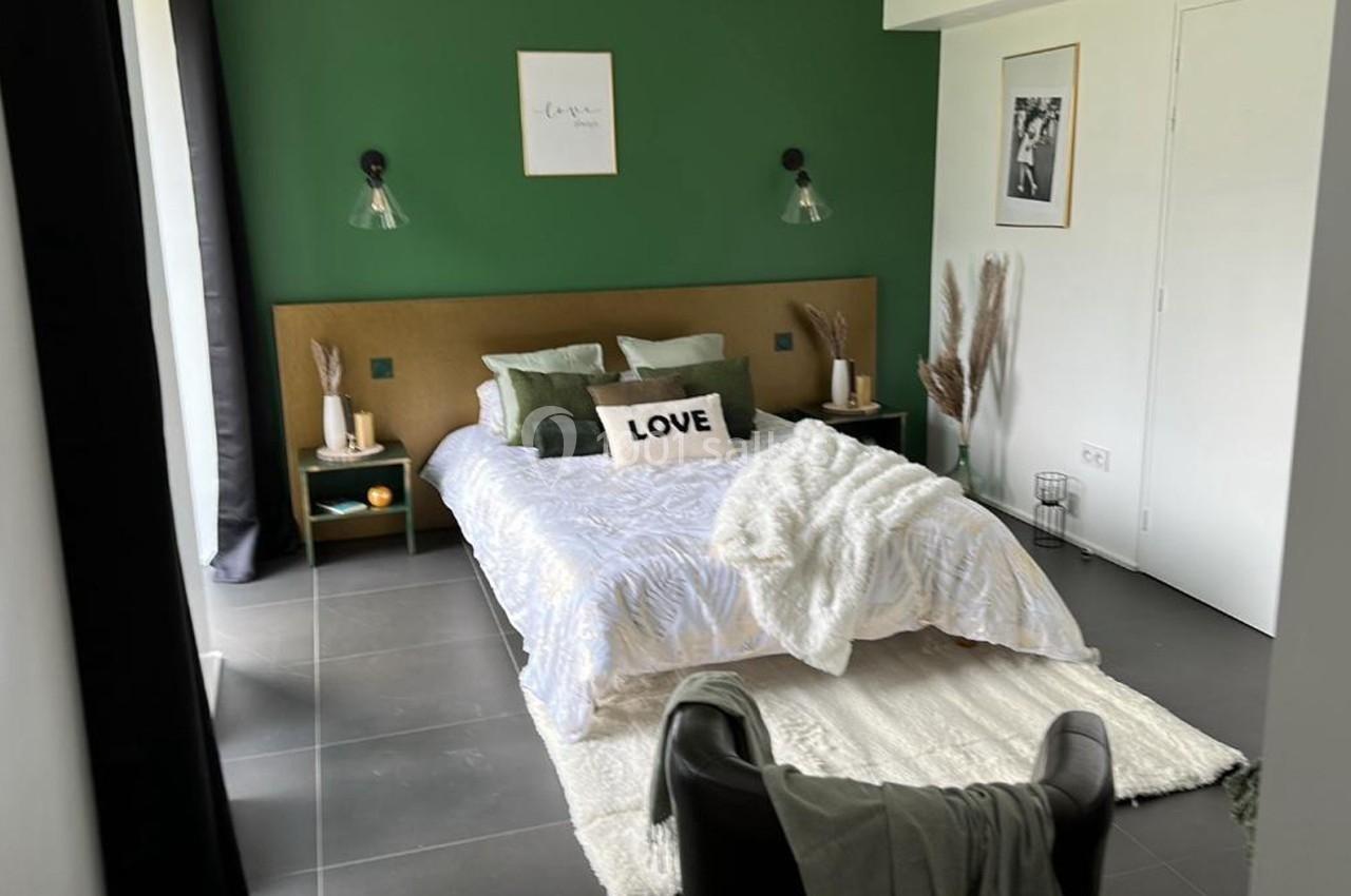 Chambre moderne avec mur vert, lit double décoré de coussins, luminaires muraux et tapis clair au sol.