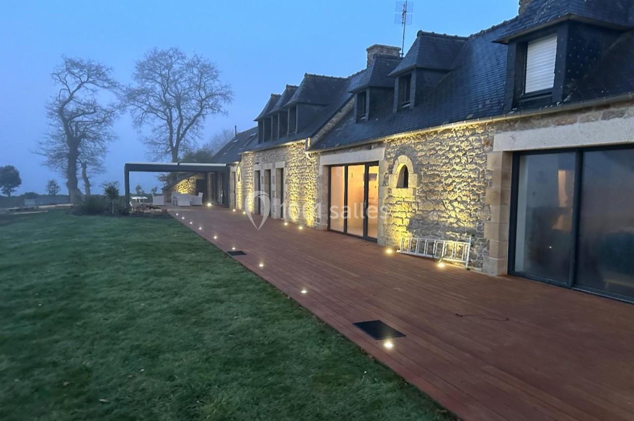 Maison en pierre avec terrasse en bois éclairée au crépuscule, entourée de pelouse et d'arbres.