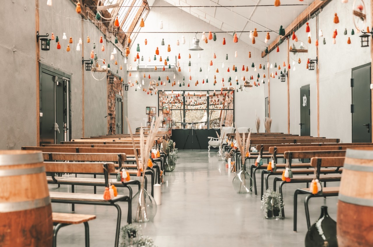 Salle décorée pour un événement, avec bancs en bois, guirlandes suspendues et fleurs dans des vases en verre.