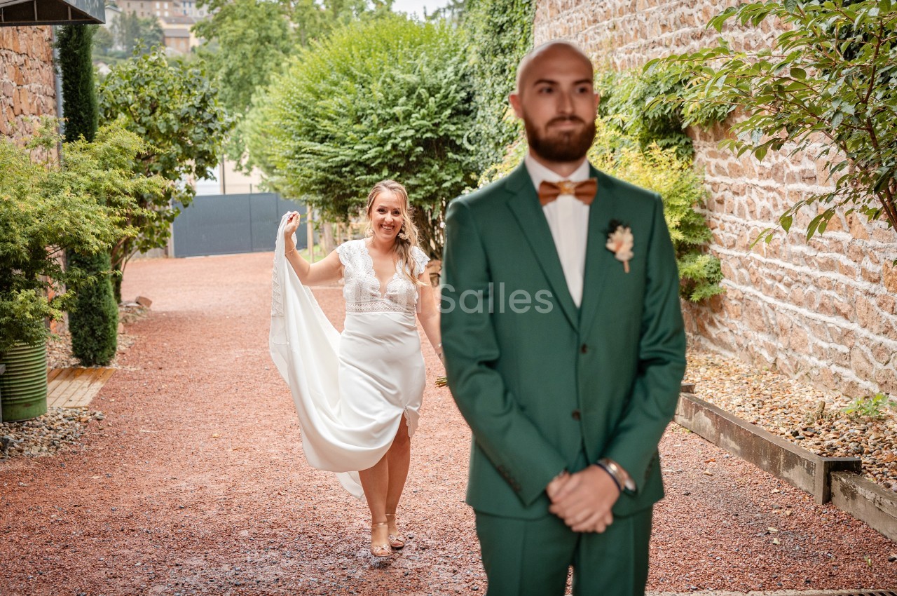 Une mariée souriante s'approche d'un marié en costume vert, debout de dos dans une allée extérieure.