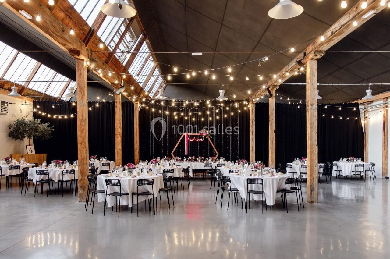 Salle de réception spacieuse avec tables décorées, guirlandes lumineuses suspendues et une arche florale en arrière-plan.