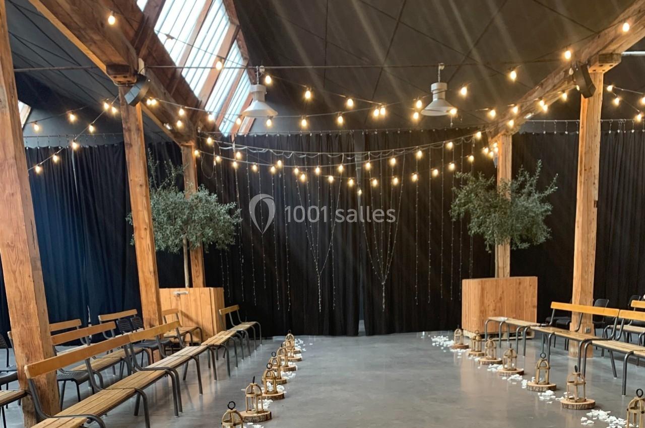 Salle décorée pour un événement avec guirlandes lumineuses suspendues, bancs en bois et lanternes alignées au sol.