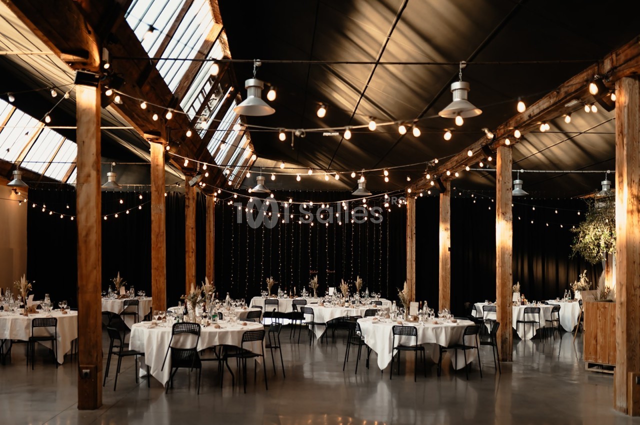 Salle de réception avec tables dressées, chaises noires, guirlandes lumineuses suspendues et poutres en bois apparentes.