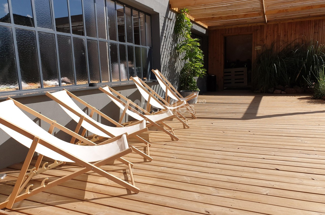 Chaises longues en bois alignées sur une terrasse en bois, près d'une grande fenêtre et d'un espace ombragé.