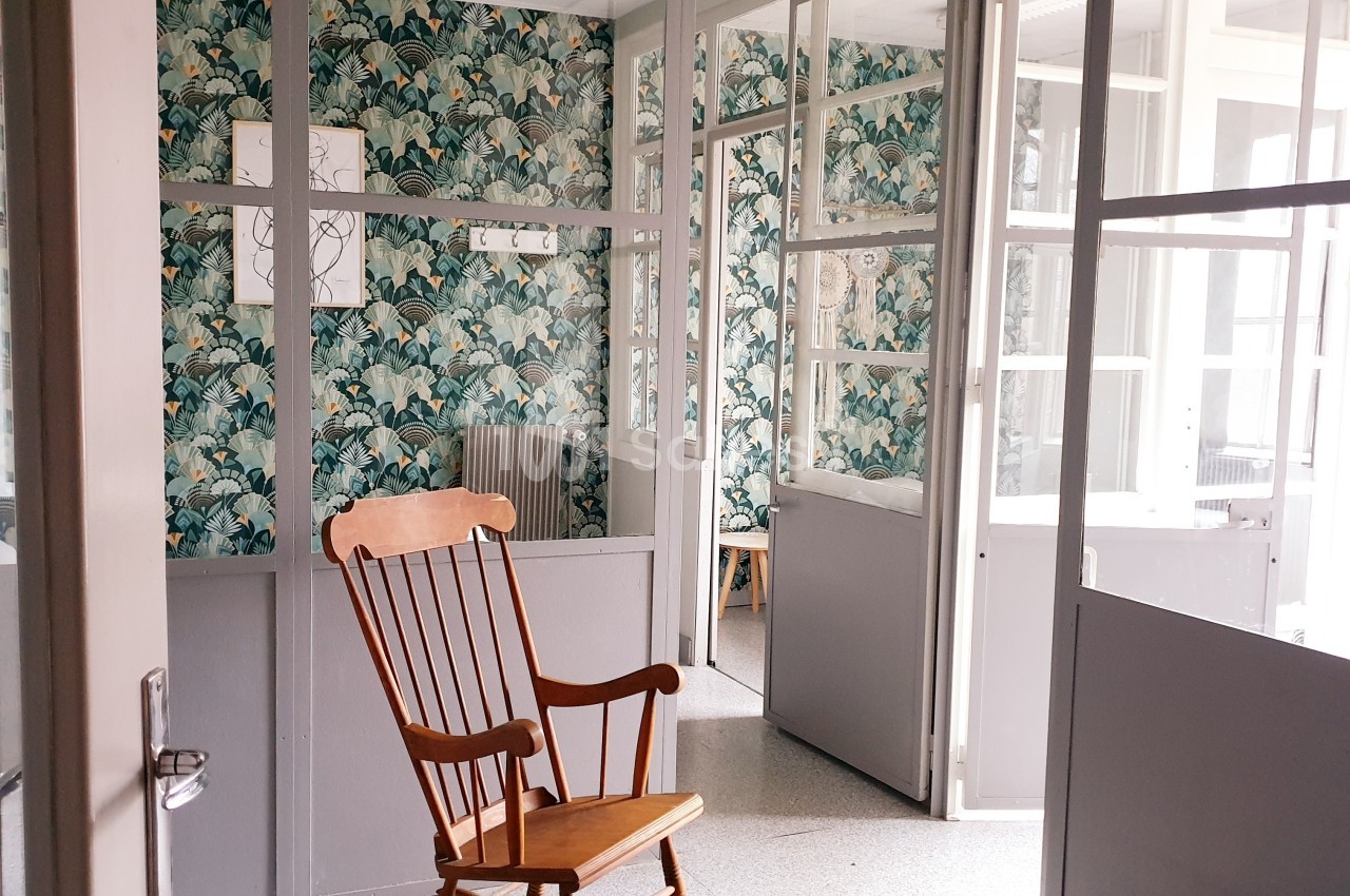 Chaise berçante en bois placée dans une pièce lumineuse avec des cloisons vitrées et un mur à motifs végétaux.