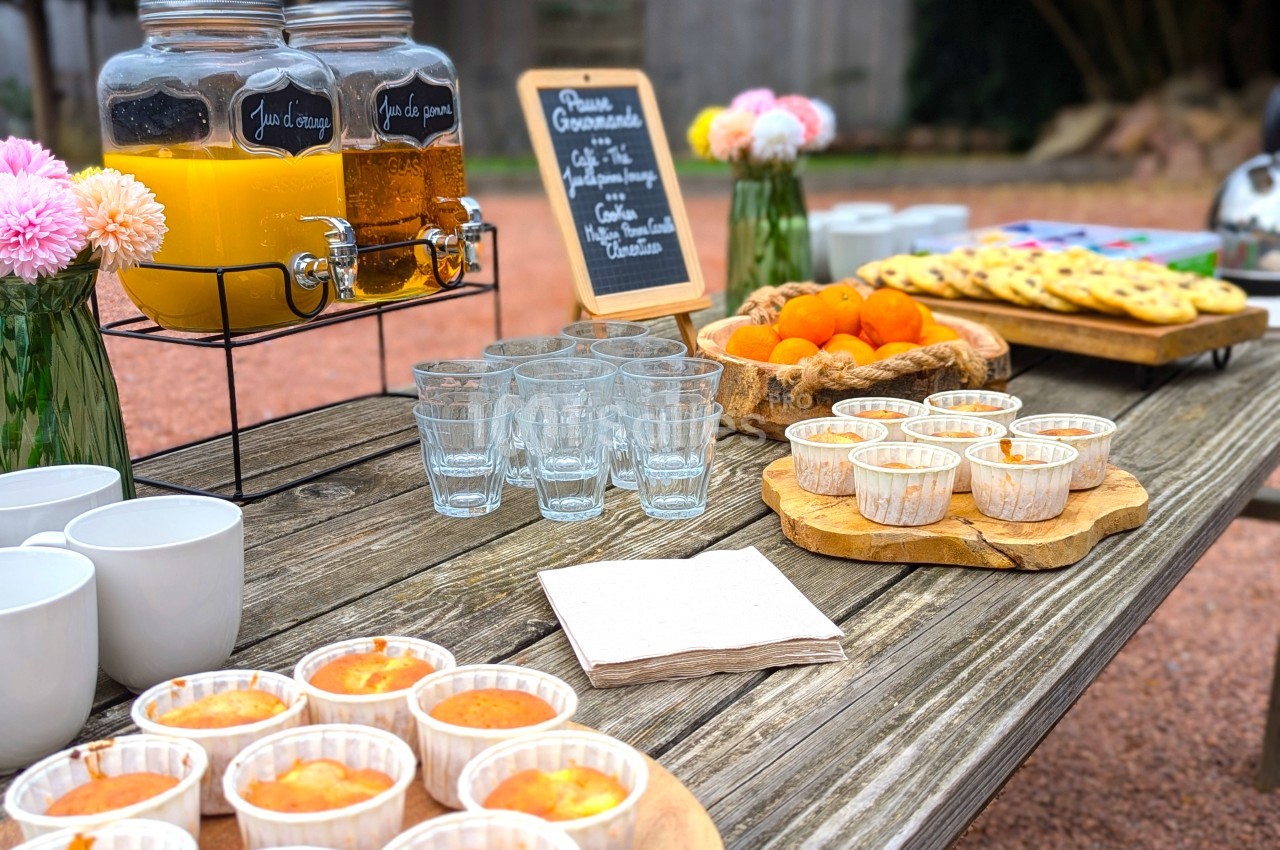 Table en bois avec boissons, verrines, fruits, biscuits et fleurs, disposés pour un buffet en extérieur.