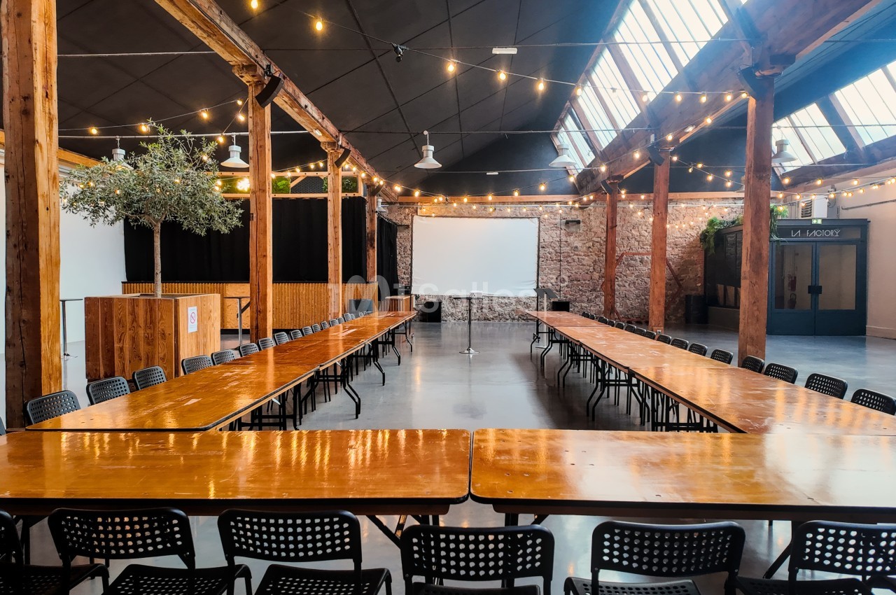 Salle spacieuse avec de longues tables en bois, chaises noires, projecteur et guirlandes lumineuses suspendues.