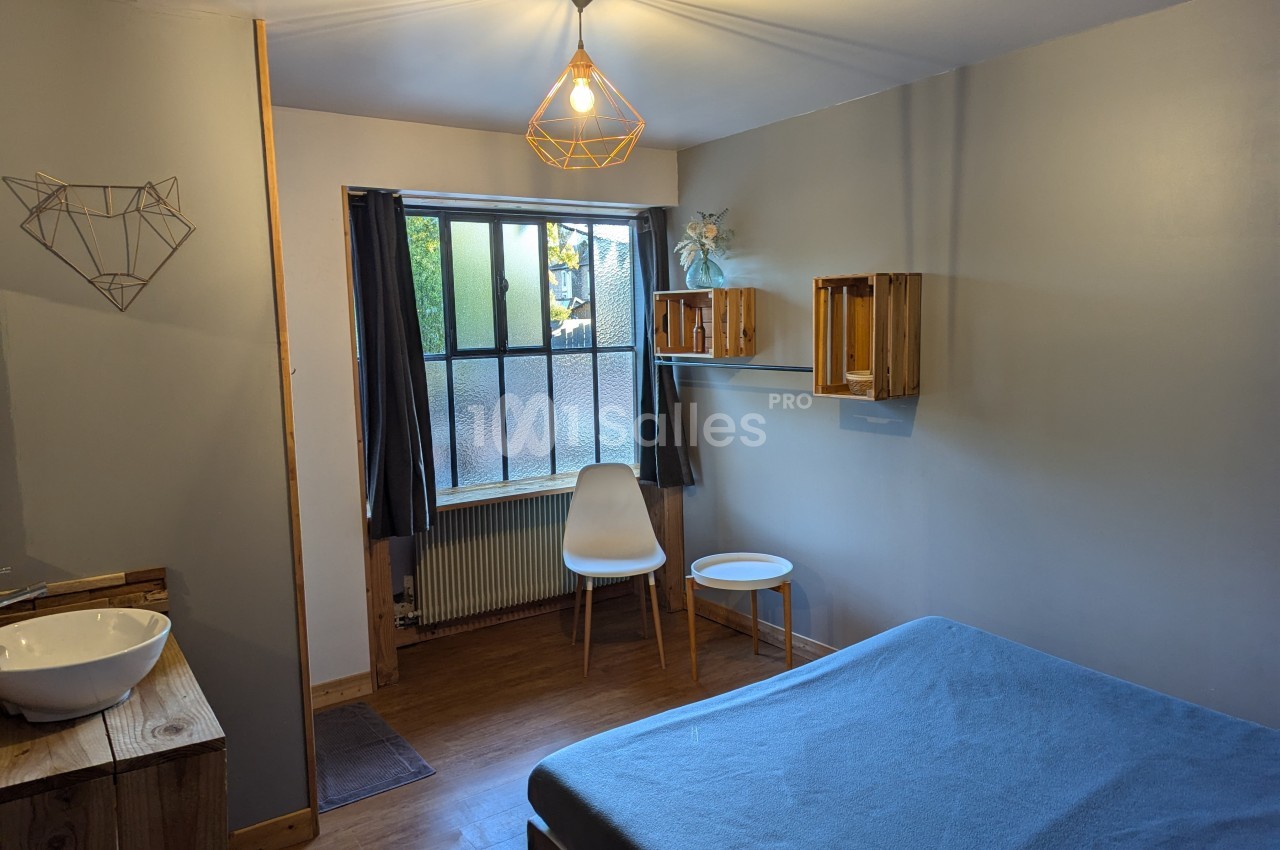 Chambre lumineuse avec lit, lavabo, chaise blanche, table basse, étagères en bois et grande fenêtre donnant sur l'extérieur.